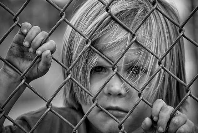 a sad child standing behind a chain link fence and holding on to the wire as he looks out beyond the fence