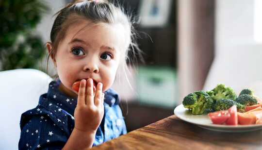 A little girl eats vegetables from a plate while sitting at a table