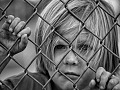 a sad child standing behind a chain link fence and holding on to the wire as he looks out beyond the fence
