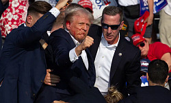 Trump with blood on his face holds up a clenched fist while people in suits surround him, illustrating the impact of social media on political violence.