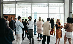 A diverse crowd of people in an office building, standing in small groups having conversations, representing networking in a professional environment.