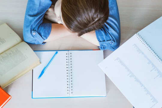 A student rests her head, sleeping on a table, with open books in front of her.