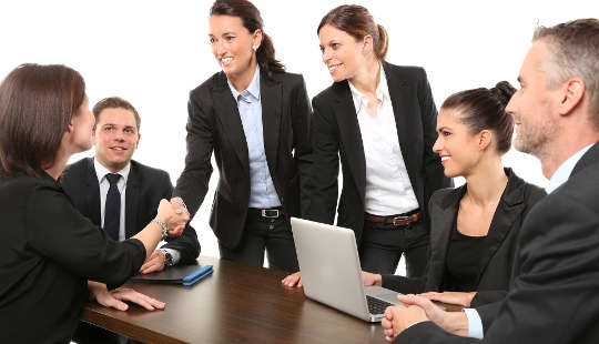 women shaking hands at a business meeting, with men loooking on