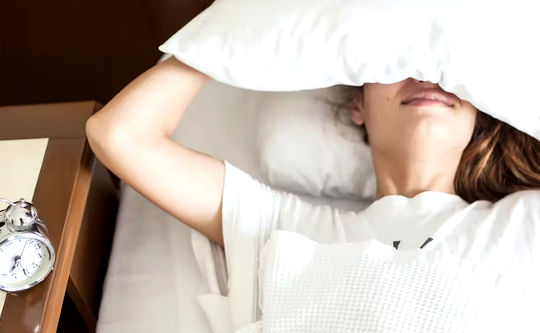 person laying in bed with pillow over their head and alarm clock next to the bed