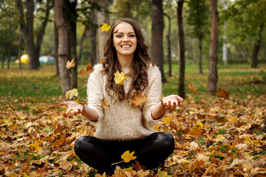 a smiling young woman sitting in the forest amidst falling autumn leaves