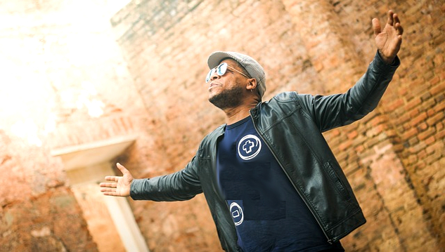 a man, wearing sunglasses, with arms wide open and looking up at the sky