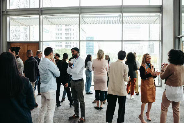 A diverse crowd of people in an office building, standing in small groups having conversations, representing networking in a professional environment.