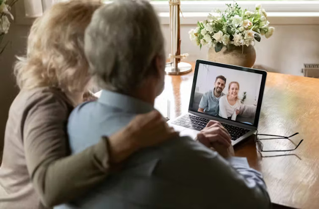 A caregiver sitting at a desk, looking at a laptop while holding a phone, symbolizing the challenges of coordinating care remotely from a long distance.