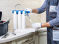 A man in work clothes installing a water filtration system under the kitchen sink, representing the process of reducing PFAS contamination in household water.