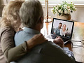 A caregiver sitting at a desk, looking at a laptop while holding a phone, symbolizing the challenges of coordinating care remotely from a long distance.