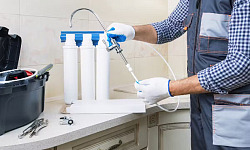 A man in work clothes installing a water filtration system under the kitchen sink, representing the process of reducing PFAS contamination in household water.