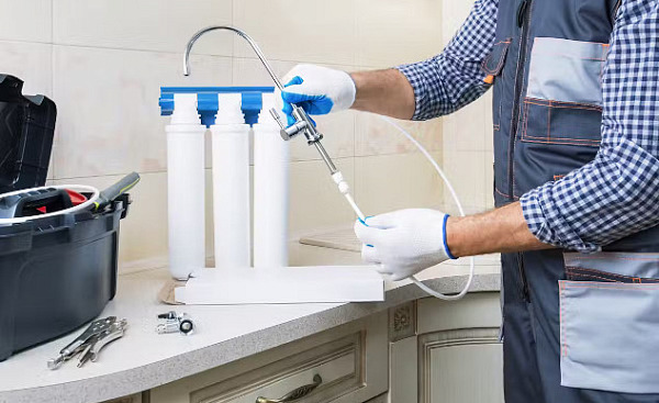 A man in work clothes installing a water filtration system under the kitchen sink, representing the process of reducing PFAS contamination in household water.