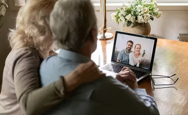 A caregiver sitting at a desk, looking at a laptop while holding a phone, symbolizing the challenges of coordinating care remotely from a long distance.