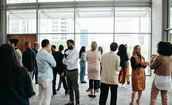 A diverse crowd of people in an office building, standing in small groups having conversations, representing networking in a professional environment.