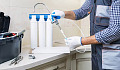 A man in work clothes installing a water filtration system under the kitchen sink, representing the process of reducing PFAS contamination in household water.