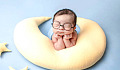 baby with eyes closed wearing huge glasses and resting on a crescent moon-shaped pillos